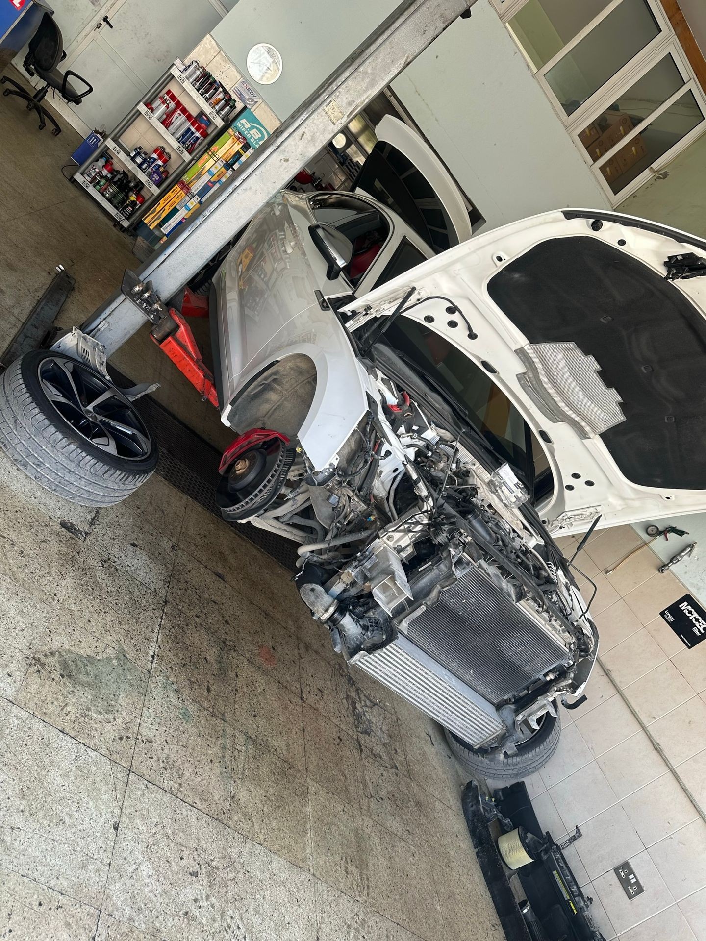 Car with hood open and front components removed, elevated on a lift in an auto repair shop.